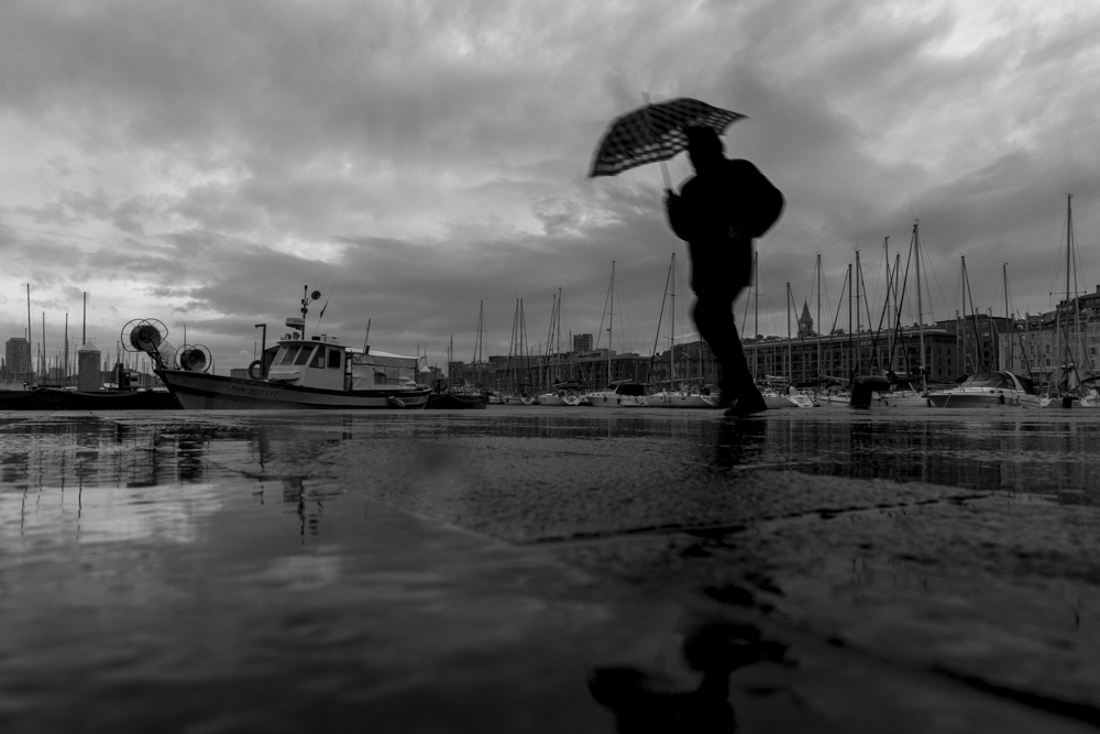 Un homme à l'abri de son parapluie presse le pas sur le Vieux-Port de Marseille, sous un ciel chargé de nuages et une pluie battante.