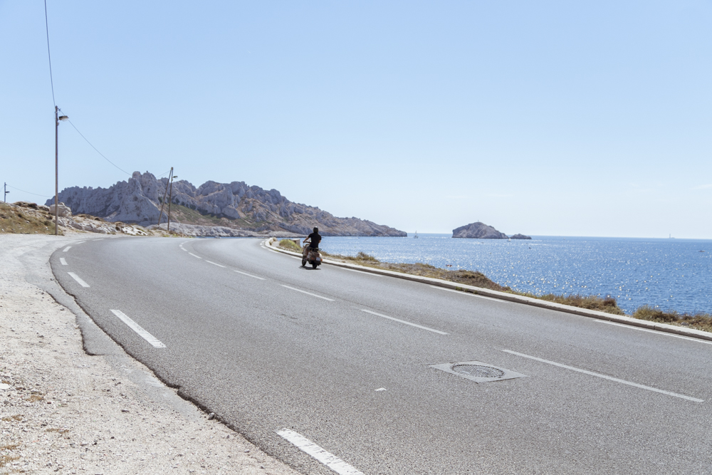 Un homme sur un scooter, sous un beau soleil d'été, suit la route menant au Bout du Monde longeant la Méditerranée.