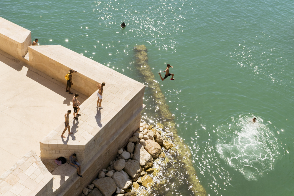 Un jeune garçon saute dans la Méditerranée à partir des remparts en contre-bas du Fort Saint-Jean sous le regard des ses camarades de jeu.