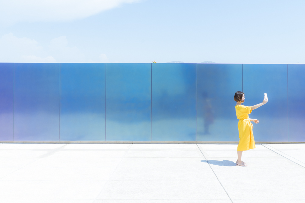 Une jeune femme vêtue d'une robe jaune, sur le toit-terrasse de la Cité Radieuse du Corbusier se prenant en photo avec son téléphone.