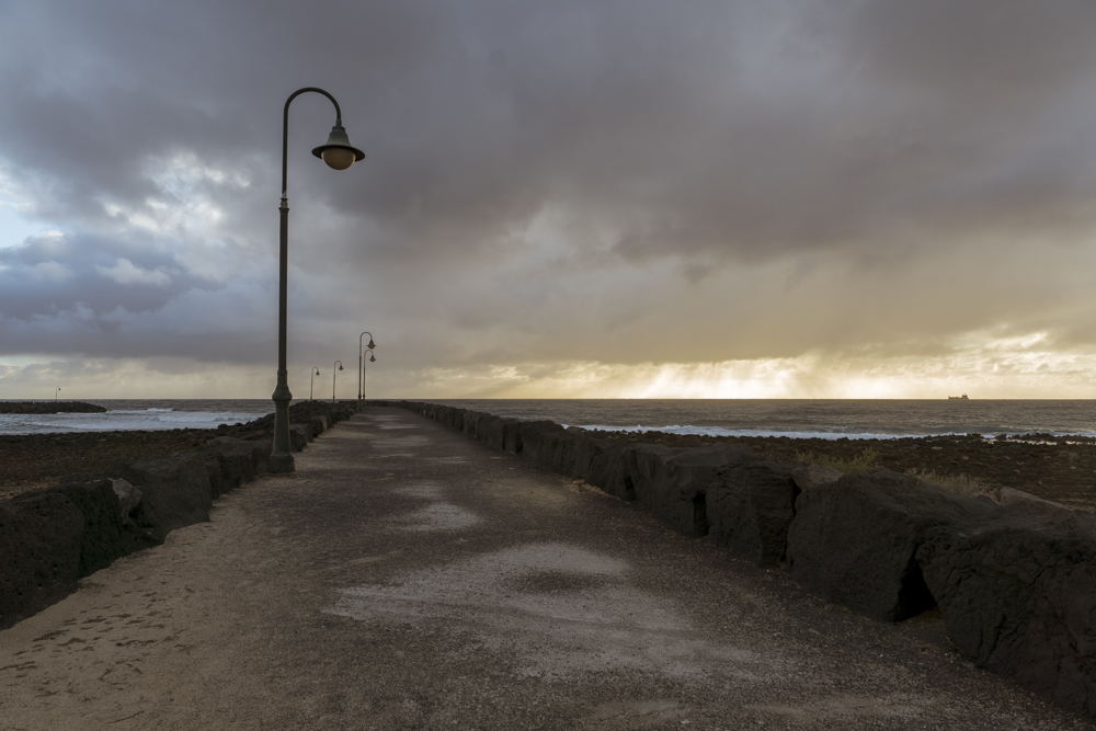 Un sentier balisé par des rochers noirs et des lampadaires, semble plonger dans l'océan Atlantique sous un ciel menaçant.
