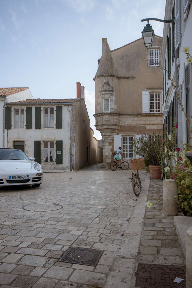 Une rue d'Ars-en-Ré en fin d'après-midi où se croise véhicule de luxe et vélo devant une ancienne bâtisse.