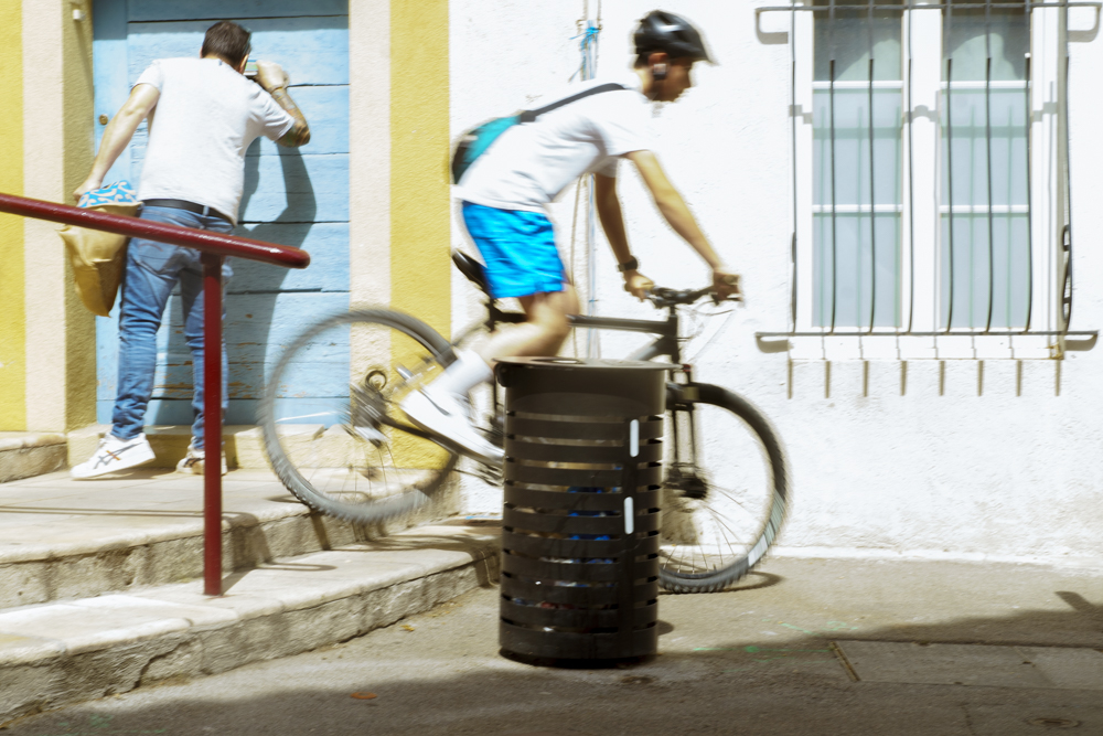 Un homme regarde par l'ouverture d'une boîte aux lettre dans une porte. Un autre sur son vélo dévale des escaliers.
