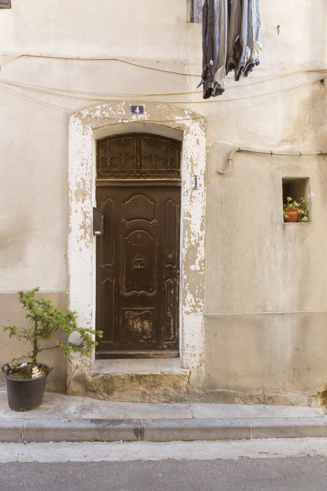 Une vieille porte en bois dans un encadrement de pierre marque l'entrée d'une maison de village. Du linge étendu à une fenêtre de l'étage ainsi qu'une plante dans un pot sur le trottoir complète la façade.