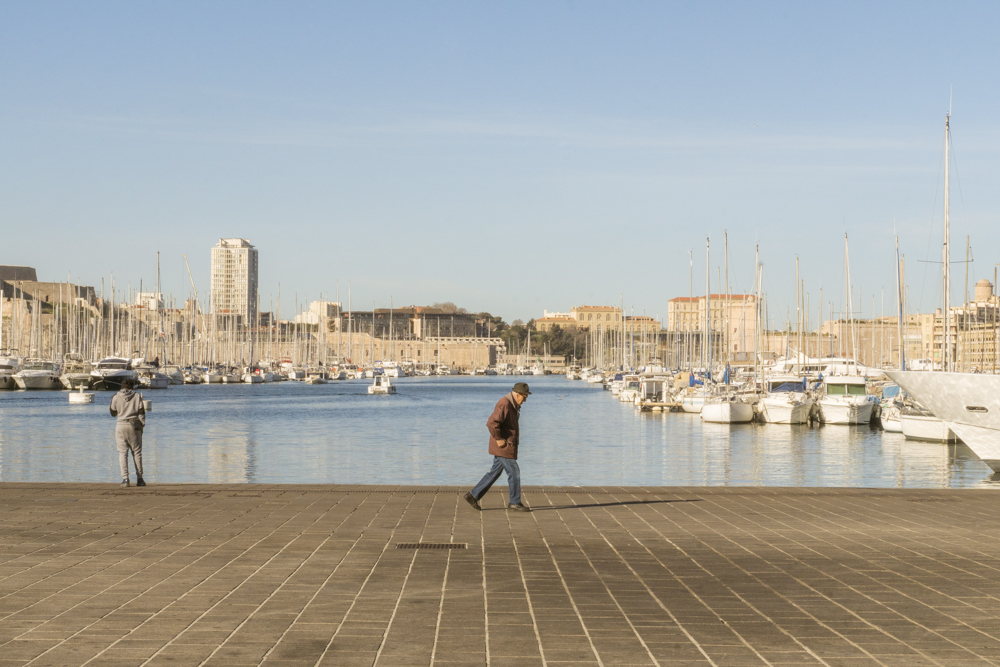 Un homme seul marche le dos courbé sur le Vieux-Port à Marseille.