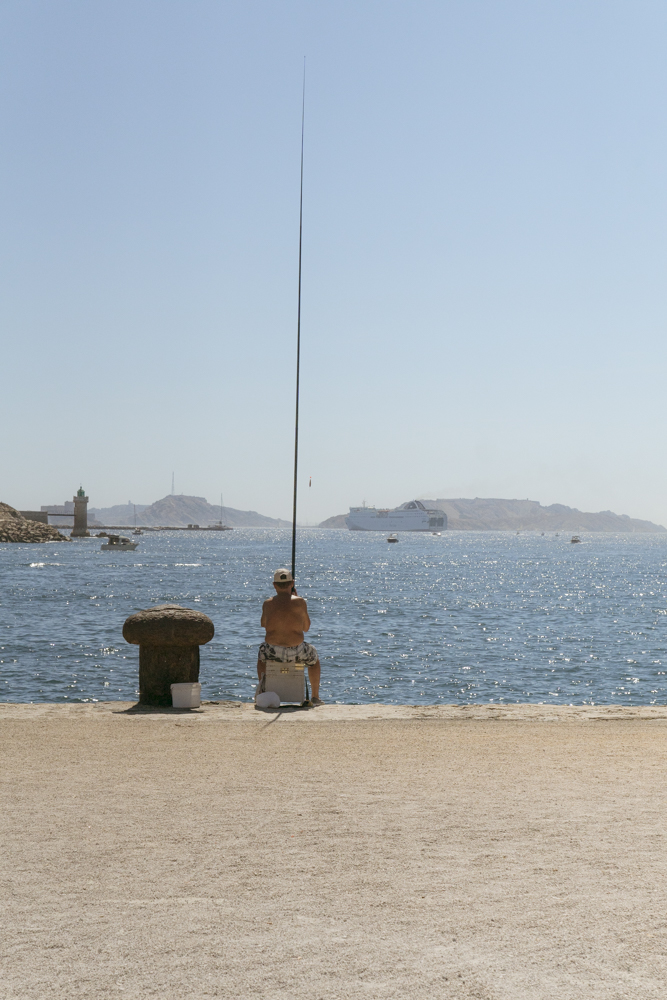 Un homme assis, torse nu, pêche à l'aide d'une grande canne à pêche sur un quai à côté d'une bite d'amarrage.