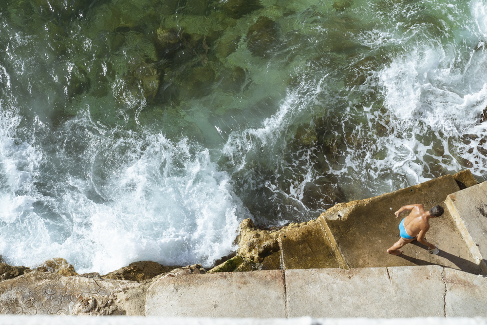 Un homme avec un petit seau en plastique remonte des rochers frappés par les vagues de la Méditerranée.