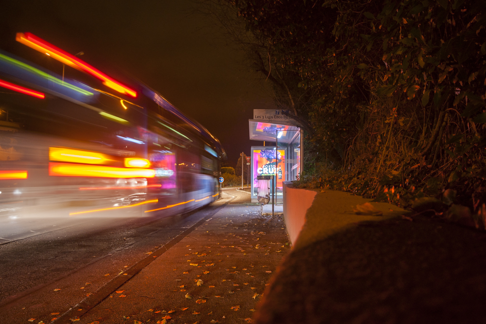 Un bus photographié en pose longue arrivant sur son arrêt semble laisser une trainée de lumière.