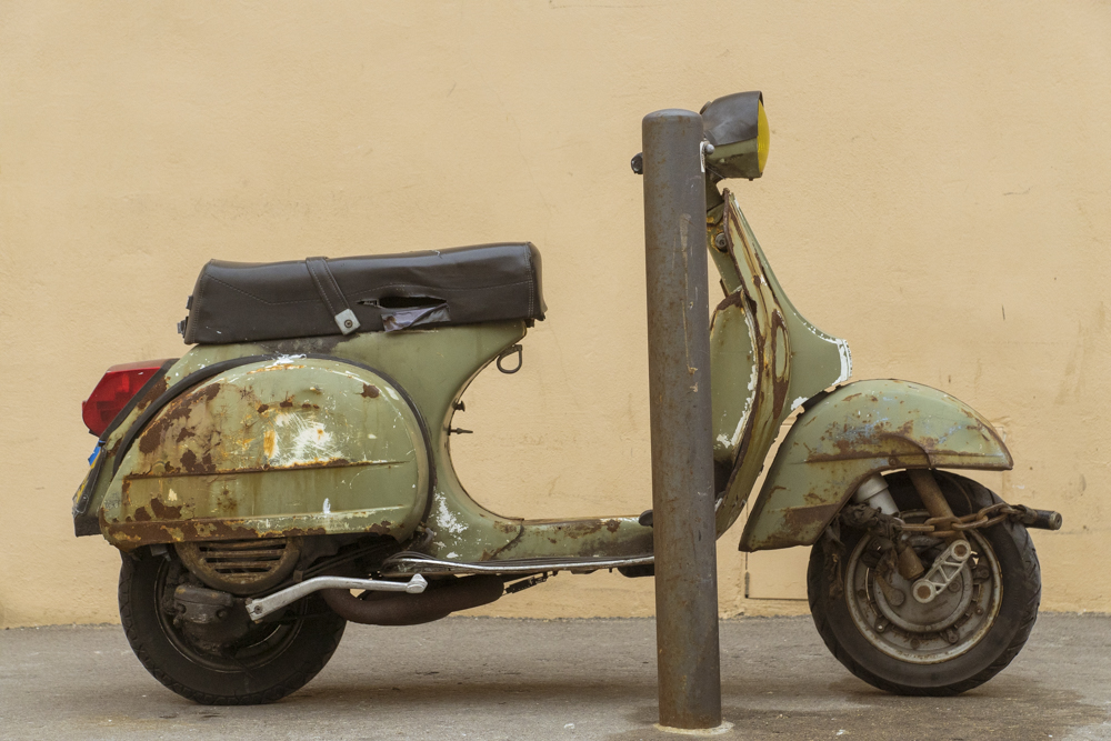 Un scooter Vespa ayant subit les outrages du temps mais qui semble ecnore en état de marche à la vue de la chaine mise à sa roue.