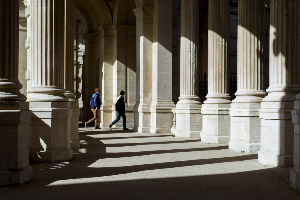 Deux hommes marchent sous les colonnades du Palais Longchamp.